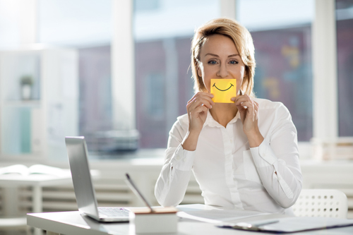 Eine Frau im Büro, die vor Ihrem Mund ein Kärtchen mit einen Smile hält.